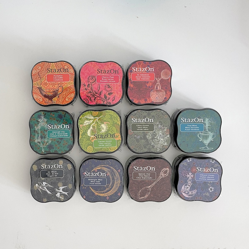 K&ouml;nnte beinhalten: Zw&ouml;lf Stempelkissen in verschiedenen Farben und Designs. Jedes Kissen hat eine andere Farbe und ein anderes Design, und jedes Kissen tr&auml;gt den Aufdruck "StazOn". Die Stempelkissen sind in einem Raster angeordnet.