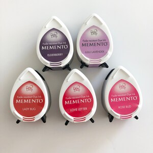 May include: Five small, teardrop-shaped ink pads with black lids. Each pad has a different color ink and a label that reads "Dew Drop Fade-resistant Dye Ink MEMENTO" followed by the ink color name: Elderberry, Lulu Lavender, Lady Bug, Love Letter, and Rose Bud.