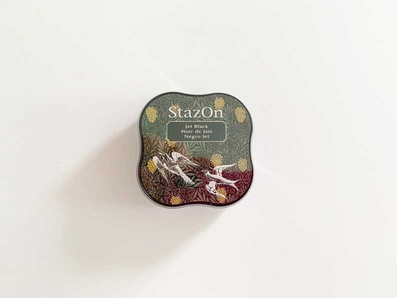 K&ouml;nnte beinhalten: Ein rundes, schwarzes Stempelkissen mit einem floralen Design und dem Text "StazOn Jet Black Noir de Jais Negro Jet".