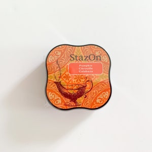 K&ouml;nnte beinhalten: Ein quadratisches, orange-braunes Stempelkissen mit einem dekorativen Design und dem Text "StazOn Pumpkin Citrouille Calabaza".