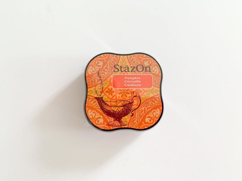 K&ouml;nnte beinhalten: Ein quadratisches, orange-braunes Stempelkissen mit einem dekorativen Design und dem Text "StazOn Pumpkin Citrouille Calabaza".
