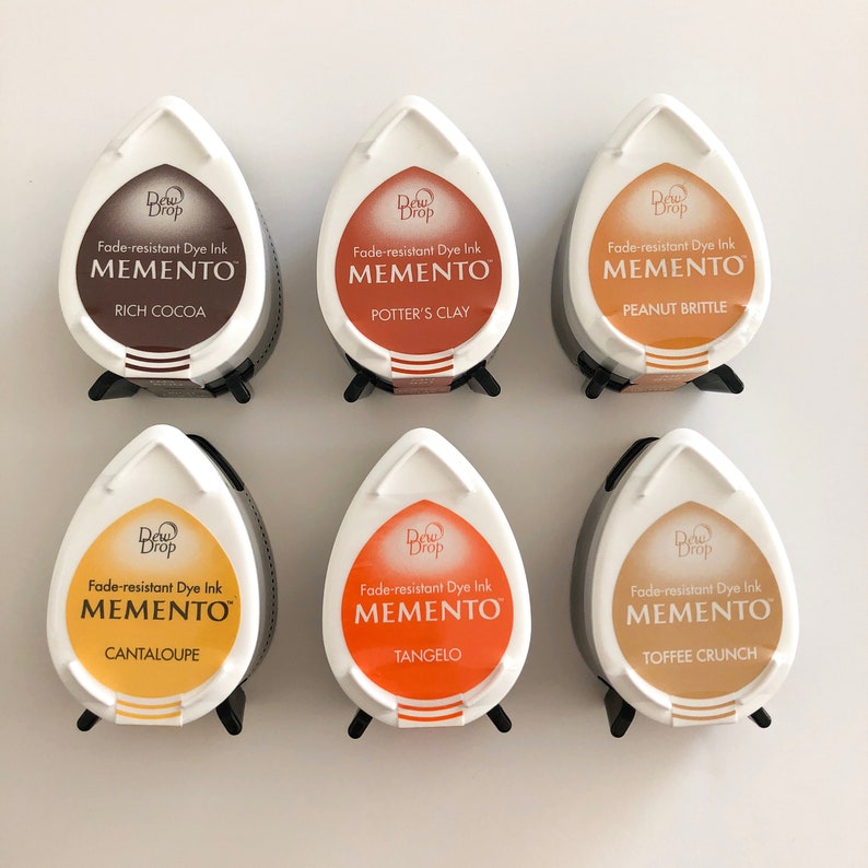 May include: Six teardrop-shaped ink pads in various shades of brown, orange, and yellow. Each pad features the text "Dew Drop Fade-resistant Dye Ink MEMENTO" and a color name, such as "Rich Cocoa" or "Toffee Crunch".