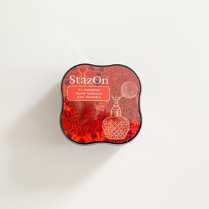 K&ouml;nnte beinhalten: Ein rotes und schwarzes Stempelkissen mit dem Text "StazOn" und "St. Valentine, Saint-Valentin, San Valentin" auf der Vorderseite. Das Stempelkissen hat ein dekoratives Design mit einer Vintage-Parf&uuml;mflasche.
