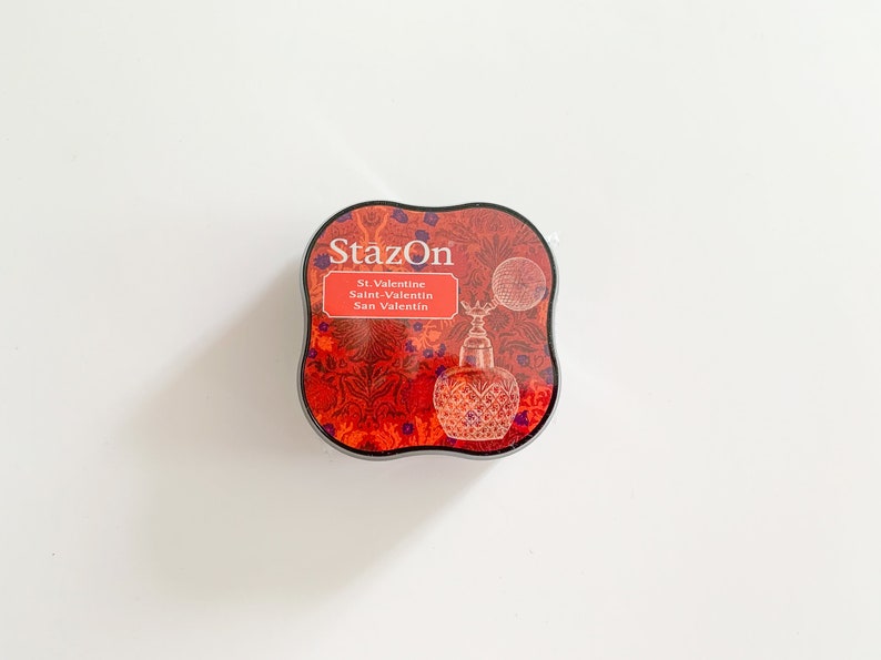 K&ouml;nnte beinhalten: Ein rotes und schwarzes Stempelkissen mit dem Text "StazOn" und "St. Valentine, Saint-Valentin, San Valentin" auf der Vorderseite. Das Stempelkissen hat ein dekoratives Design mit einer Vintage-Parf&uuml;mflasche.