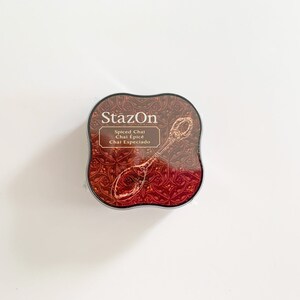K&ouml;nnte beinhalten: Ein rundes, schwarzes Stempelkissen mit einem roten und goldenen Design. Das Stempelkissen ist mit "StazOn" und "Spiced Chai Chai &Eacute;pic&eacute; Chai Especiado" beschriftet.