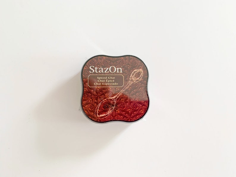 K&ouml;nnte beinhalten: Ein rundes, schwarzes Stempelkissen mit einem roten und goldenen Design. Das Stempelkissen ist mit "StazOn" und "Spiced Chai Chai &Eacute;pic&eacute; Chai Especiado" beschriftet.