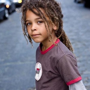 May include: A young person with dreadlocks wearing a dark brown t-shirt with red trim around the neck and sleeves. The shirt has a white patch with a red accent. The person is standing on a cobblestone street.