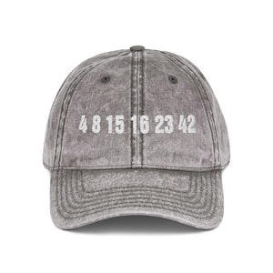 May include: Grey baseball cap featuring the numbers 4 8 15 16 23 42 in white. The cap has a curved peak and a button on top. The fabric has a worn, vintage appearance.