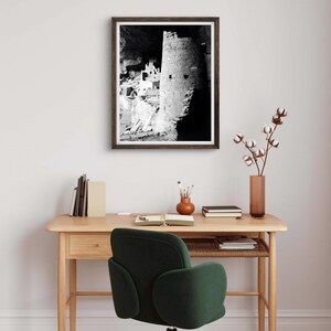 Puede incluir: Fotograf&iacute;a en blanco y negro de una vivienda en un acantilado en una cueva. La foto est&aacute; enmarcada en un marco de madera oscura y est&aacute; colgada en una pared blanca sobre un escritorio de madera con una silla verde.