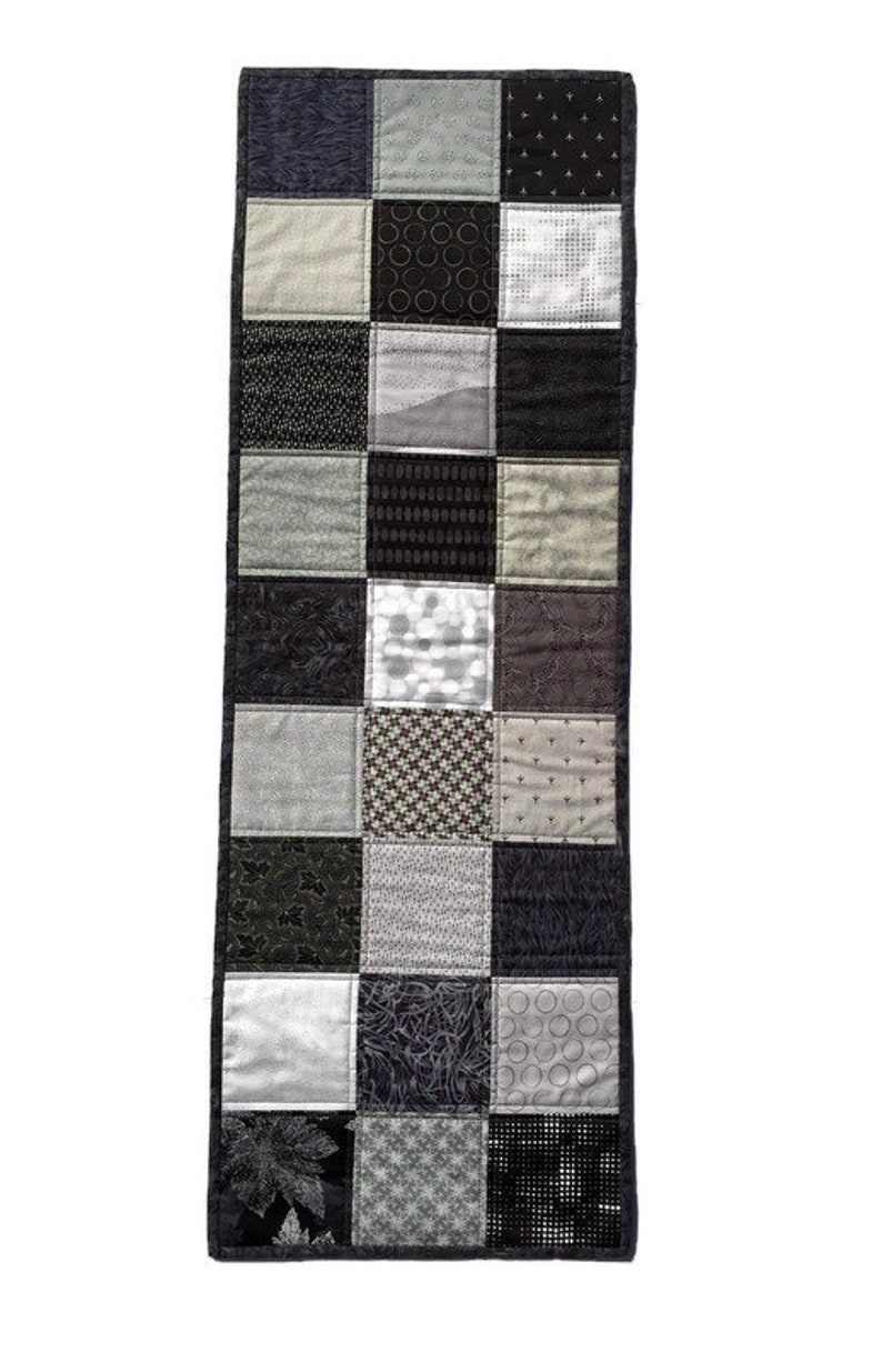 May include: A black and white patchwork table runner with a variety of geometric and floral patterns. The runner is made of fabric squares sewn together.