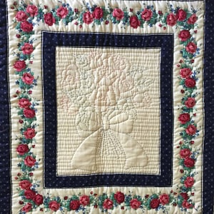 May include: A white quilted wall hanging with a floral design in the center and a border of red roses on a cream background. The border is framed by a dark blue fabric with a white polka dot pattern.