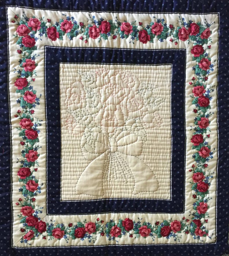 May include: A white quilted wall hanging with a floral design in the center and a border of red roses on a cream background. The border is framed by a dark blue fabric with a white polka dot pattern.