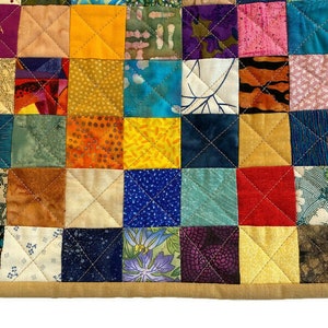 May include: A colourful patchwork quilt featuring a variety of fabrics and patterns. The quilt is made up of small squares, each with a different design. The quilt is edged with a tan fabric.
