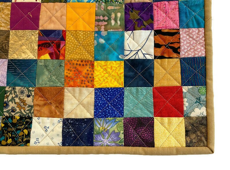 May include: A colourful patchwork quilt featuring a variety of fabrics and patterns. The quilt is made up of small squares, each with a different design. The quilt is edged with a tan fabric.