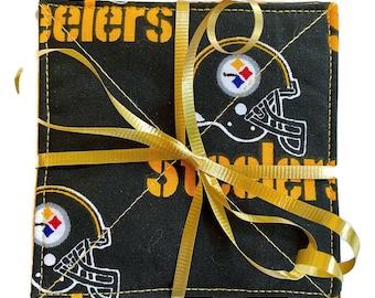 May include: Set of four black and yellow fabric coasters featuring the Pittsburgh Steelers logo and the word "Steelers" repeated on each coaster. The coasters are tied together with a gold ribbon.