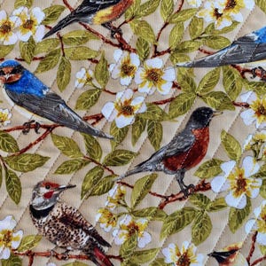 May include: A beige fabric with a quilted diamond pattern features a variety of colorful birds perched on branches with white flowers. The birds include a blue jay, a robin, a woodpecker, and a yellow bird.