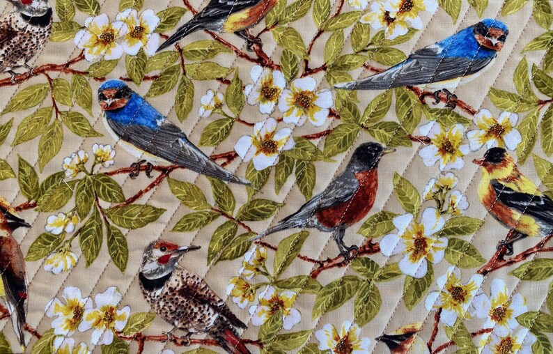 May include: A beige fabric with a quilted diamond pattern features a variety of colorful birds perched on branches with white flowers. The birds include a blue jay, a robin, a woodpecker, and a yellow bird.