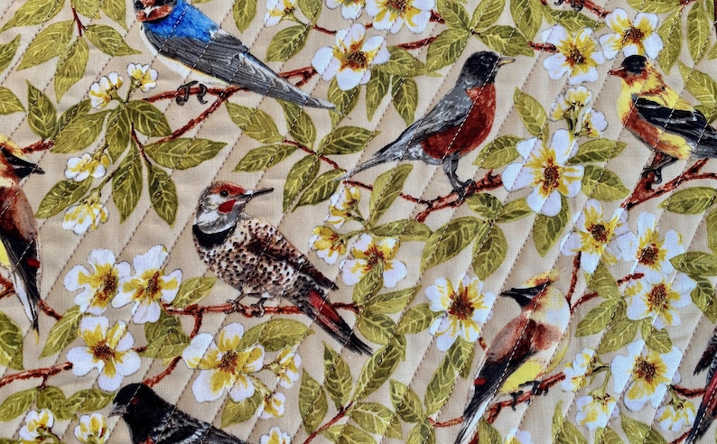 May include: A beige fabric with a quilted pattern features a variety of colorful birds and white flowers. The birds include a blue jay, a robin, a woodpecker, and a yellow bird.