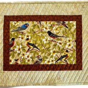 May include: A beige quilted table runner with a brown border and a central panel featuring a floral design with birds. The birds are a variety of colors, including blue, yellow, and red.