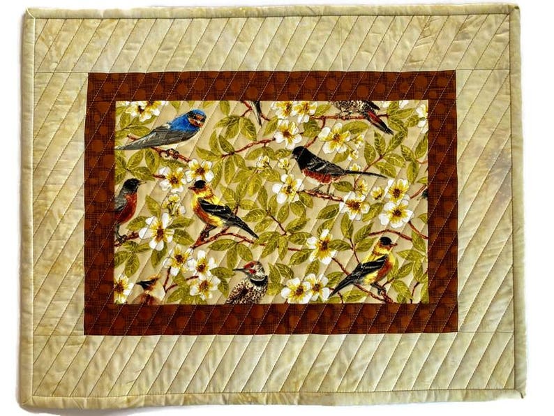 May include: A beige quilted table runner with a brown border and a central panel featuring a floral design with birds. The birds are a variety of colors, including blue, yellow, and red.