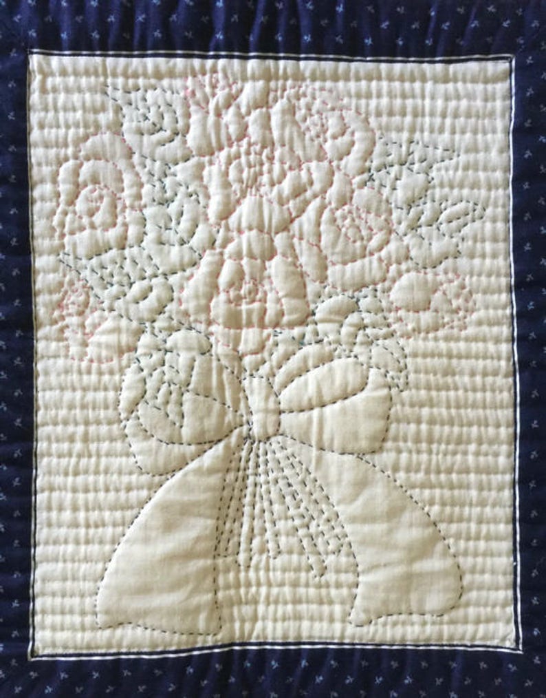 May include: A white quilted fabric square with a design of a bouquet of pink roses tied with a bow. The fabric is surrounded by a dark blue border with a small white pattern.