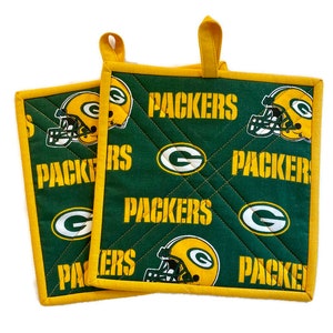May include: Two green and yellow Packers pot holders. The pot holders have a repeating pattern of the team's logo, a yellow helmet, and the word "PACKERS" in yellow. The edges are trimmed with yellow fabric and have a yellow loop for hanging.