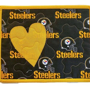May include: A quilted, rectangular trivet featuring a black background with yellow "Steelers" text and helmet graphics. A large, yellow heart is appliqued on the front. The trivet has a yellow border.
