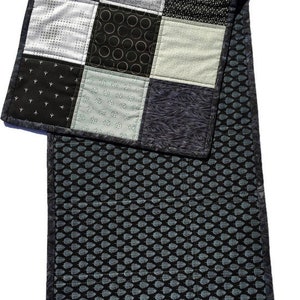 May include: A black and white patchwork table runner with a geometric pattern. The runner is made of different fabrics with various textures and patterns. The runner is quilted and has a black and white leaf pattern on the back.