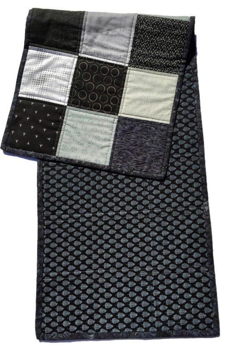May include: A black and white patchwork table runner with a geometric pattern. The runner is made of different fabrics with various textures and patterns. The runner is quilted and has a black and white leaf pattern on the back.