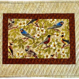 May include: A quilted placemat with a brown border and a beige background featuring a colorful floral pattern with birds.