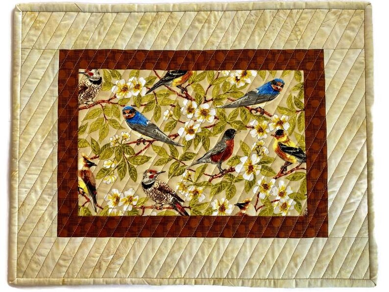 May include: A quilted placemat with a brown border and a beige background featuring a colorful floral pattern with birds.