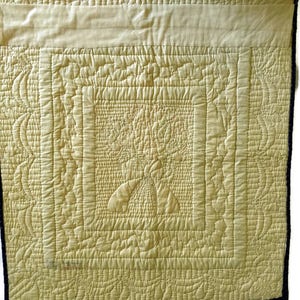 May include: A cream-colored quilted fabric with a detailed pattern of stitching. The fabric is framed with a dark blue border.
