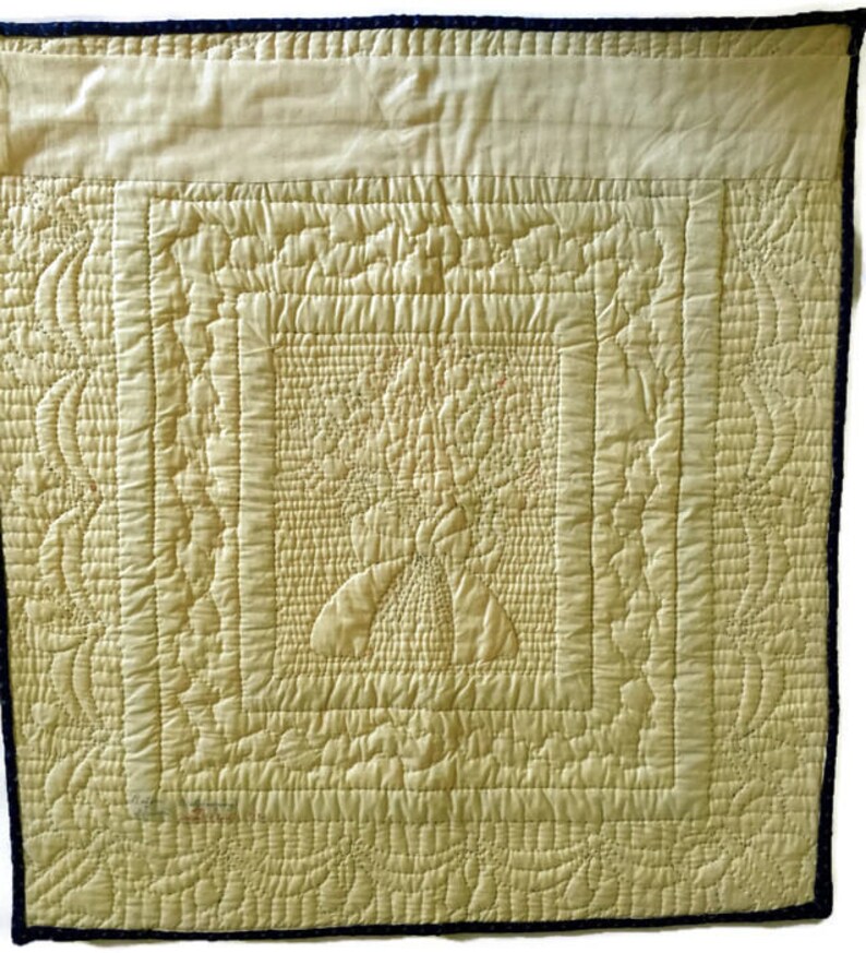 May include: A cream-colored quilted fabric with a detailed pattern of stitching. The fabric is framed with a dark blue border.