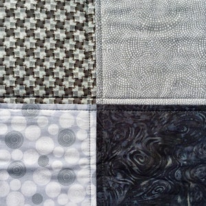 May include: A patchwork quilt with four different fabric patterns. The top left square is a gray and white houndstooth pattern. The top right square is a gray and white polka dot pattern. The bottom left square is a gray and white circle pattern. The bottom right square is a dark gray and black swirl pattern.