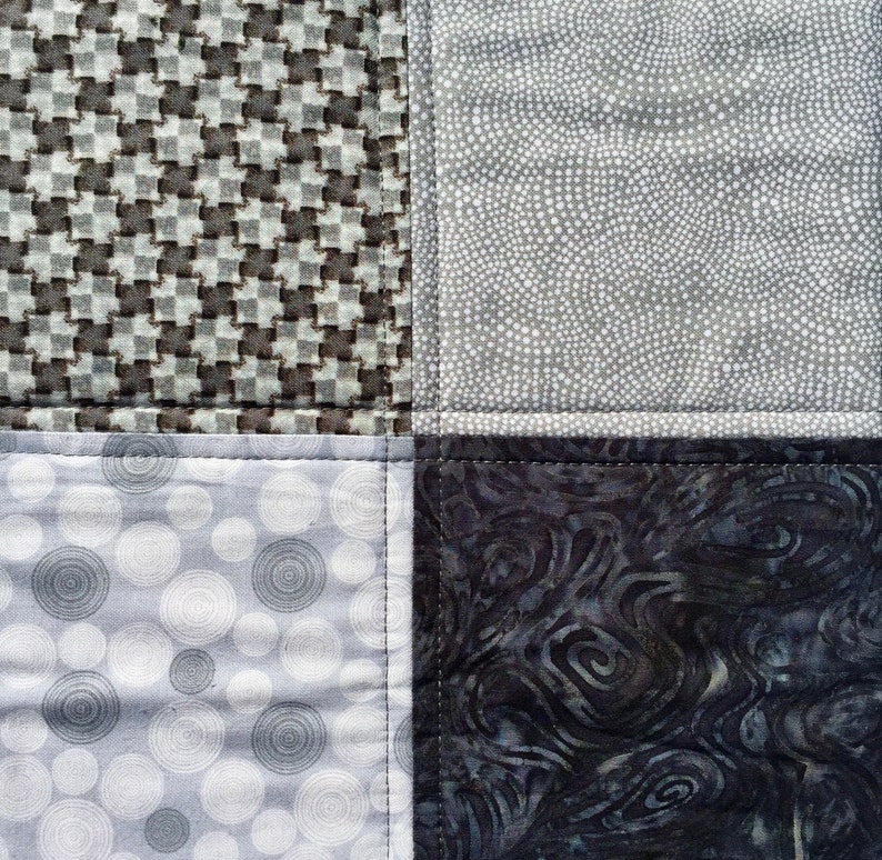May include: A patchwork quilt with four different fabric patterns. The top left square is a gray and white houndstooth pattern. The top right square is a gray and white polka dot pattern. The bottom left square is a gray and white circle pattern. The bottom right square is a dark gray and black swirl pattern.