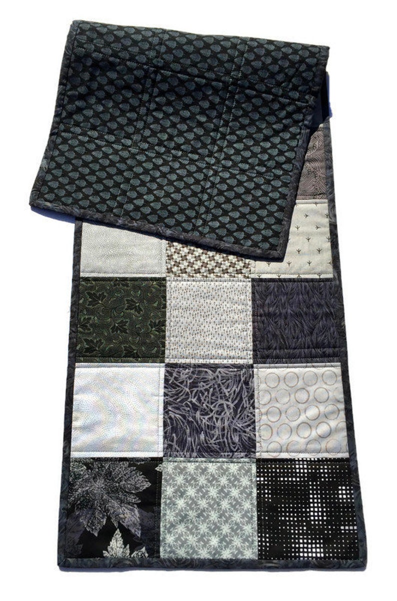 May include: A black and white patchwork table runner with a geometric pattern. The runner is made of different fabrics with various patterns, including leaves, circles, and stars.