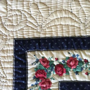 May include: Close-up of a white quilt with a floral pattern. The quilt is quilted with a design of curved lines and circles. The quilt is edged with a blue and white border.