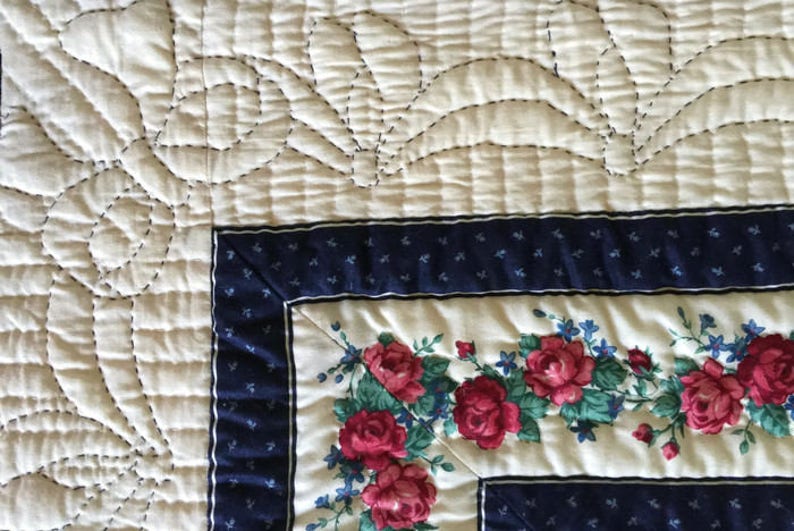 May include: Close-up of a white quilt with a floral pattern. The quilt is quilted with a design of curved lines and circles. The quilt is edged with a blue and white border.