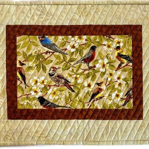 May include: A quilted table runner with a brown border and a central panel featuring a floral design with birds. The birds are in various colors, including blue, red, yellow, and brown.