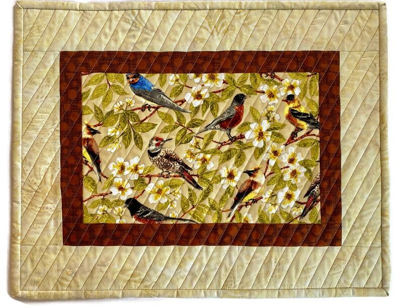 May include: A quilted table runner with a brown border and a central panel featuring a floral design with birds. The birds are in various colors, including blue, red, yellow, and brown.