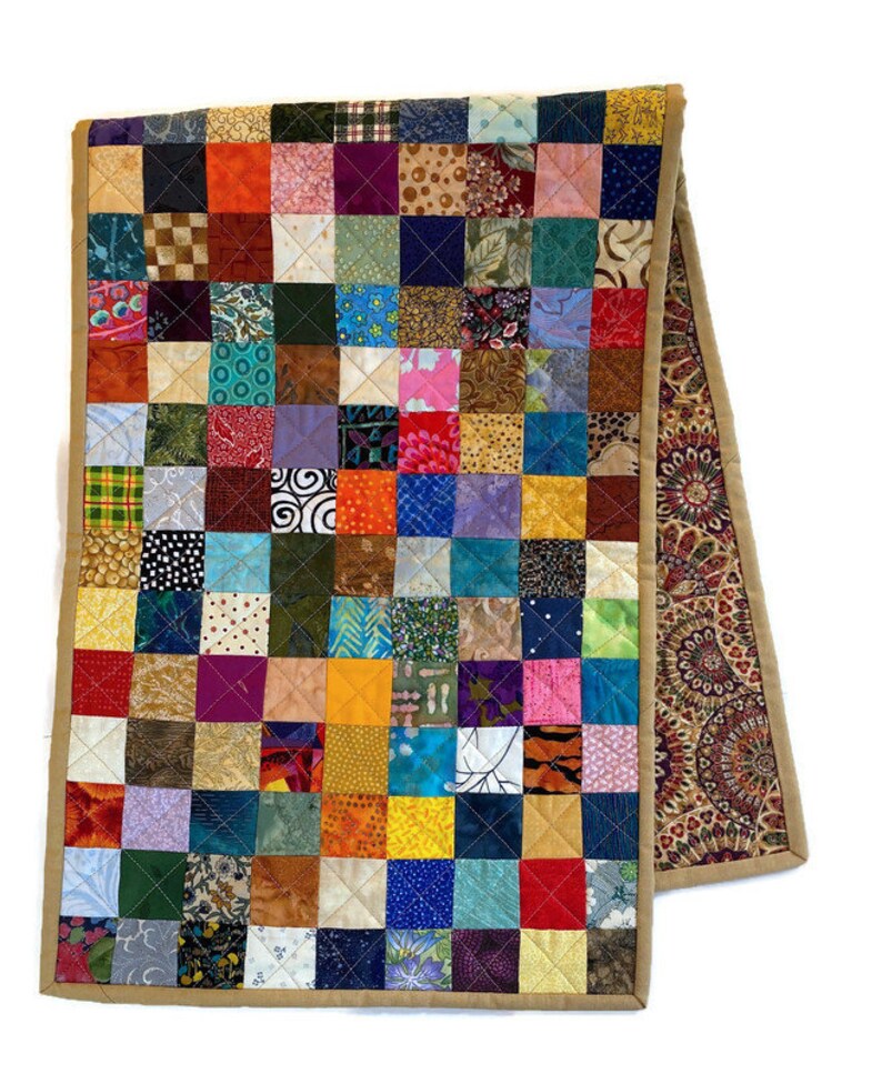May include: A colourful patchwork table runner with a tan border. The runner is made of many different fabrics, each with a unique pattern. The runner is approximately 35 cm wide and 183 cm long.