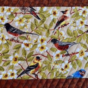May include: A beige fabric with a quilted pattern features a repeating design of white flowers, green leaves, and colorful birds. The birds are depicted in various poses, including perched on branches and flying.