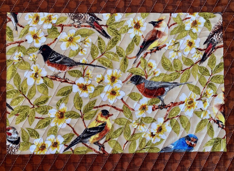 May include: A beige fabric with a quilted pattern features a repeating design of white flowers, green leaves, and colorful birds. The birds are depicted in various poses, including perched on branches and flying.