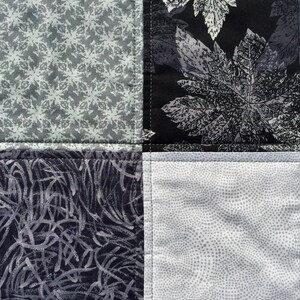 May include: A patchwork quilt with four squares. The top left square is a gray and white geometric pattern. The top right square is a black and gray floral pattern. The bottom left square is a black and gray abstract pattern. The bottom right square is a white and gray polka dot pattern.