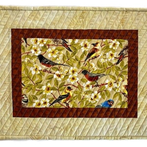 May include: A quilted placemat with a brown border and a central design of birds and flowers. The birds are in various colors, including blue, red, yellow, and brown. The flowers are white and yellow.
