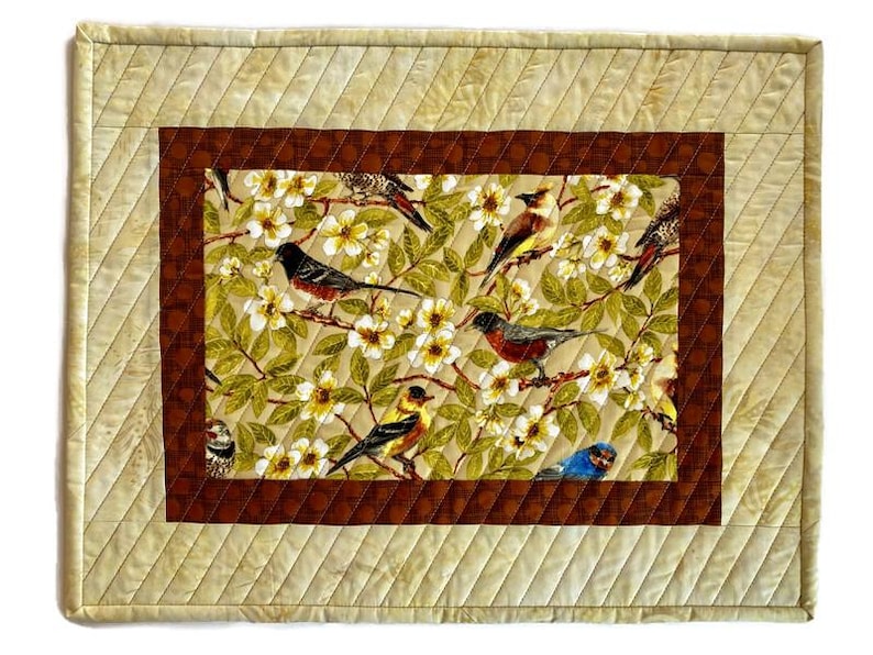 May include: A quilted placemat with a brown border and a central design of birds and flowers. The birds are in various colors, including blue, red, yellow, and brown. The flowers are white and yellow.