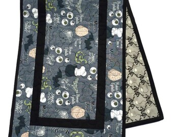 May include: A gray and black quilted table runner with a spooky Halloween theme. The fabric features a repeating pattern of eyeballs, brains, bats, snakes, and the text "Moth Dust", "Snake & Oil", and "Brew".
