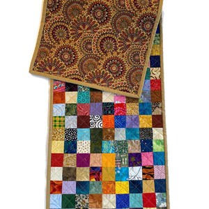 May include: A colourful patchwork table runner with a brown and gold patterned fabric on the back. The runner is made of squares of different fabrics in a variety of colours and patterns.