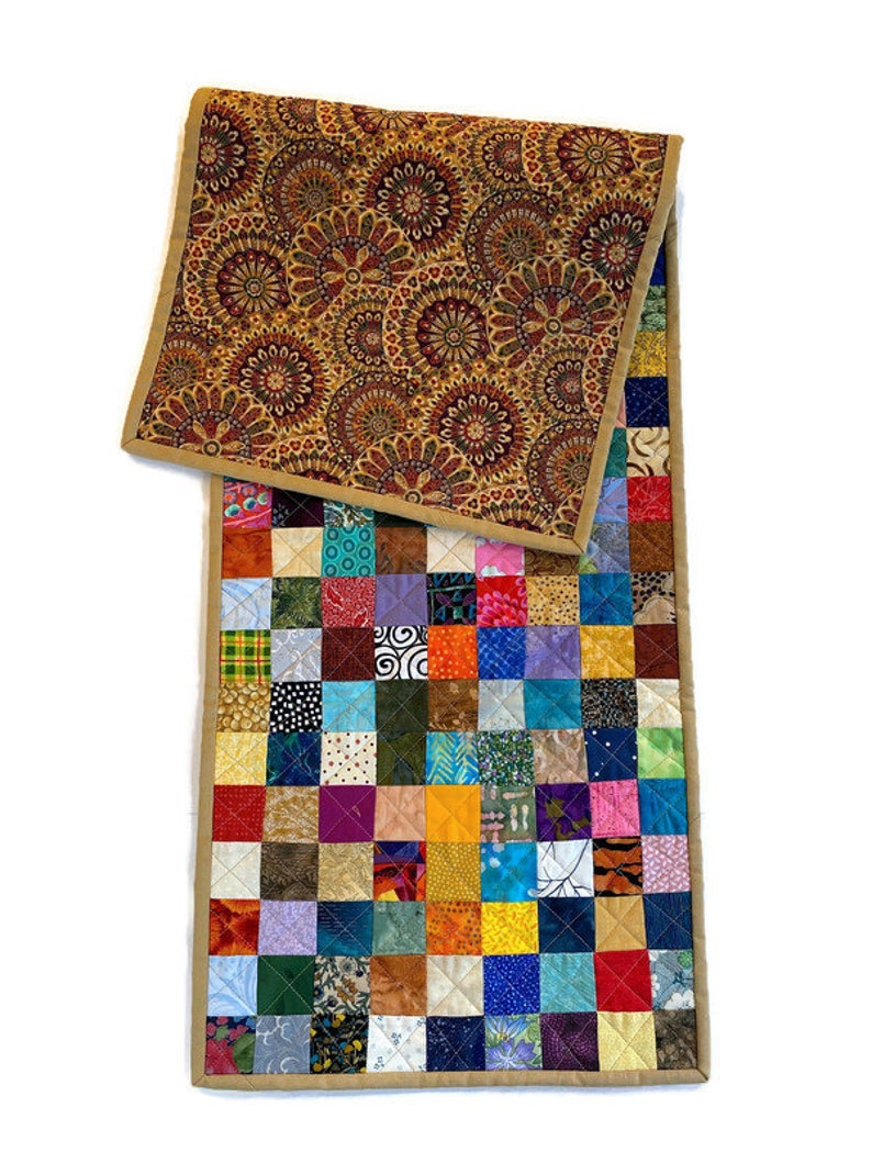 May include: A colourful patchwork table runner with a brown and gold patterned fabric on the back. The runner is made of squares of different fabrics in a variety of colours and patterns.