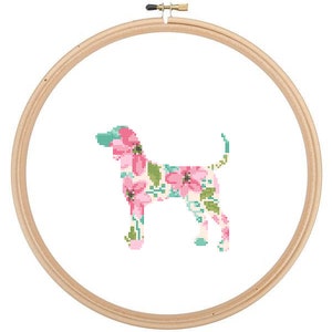 以下が含まれることがあります： 木製の刺繍枠の中に、犬のフローラルパターンがクロスステッチでデザインされています。